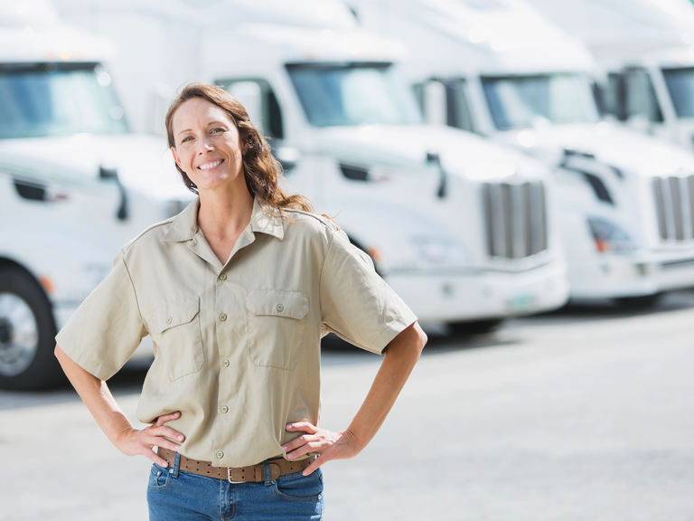 A woman smiles, standing in front of a row of parked semi trucks.